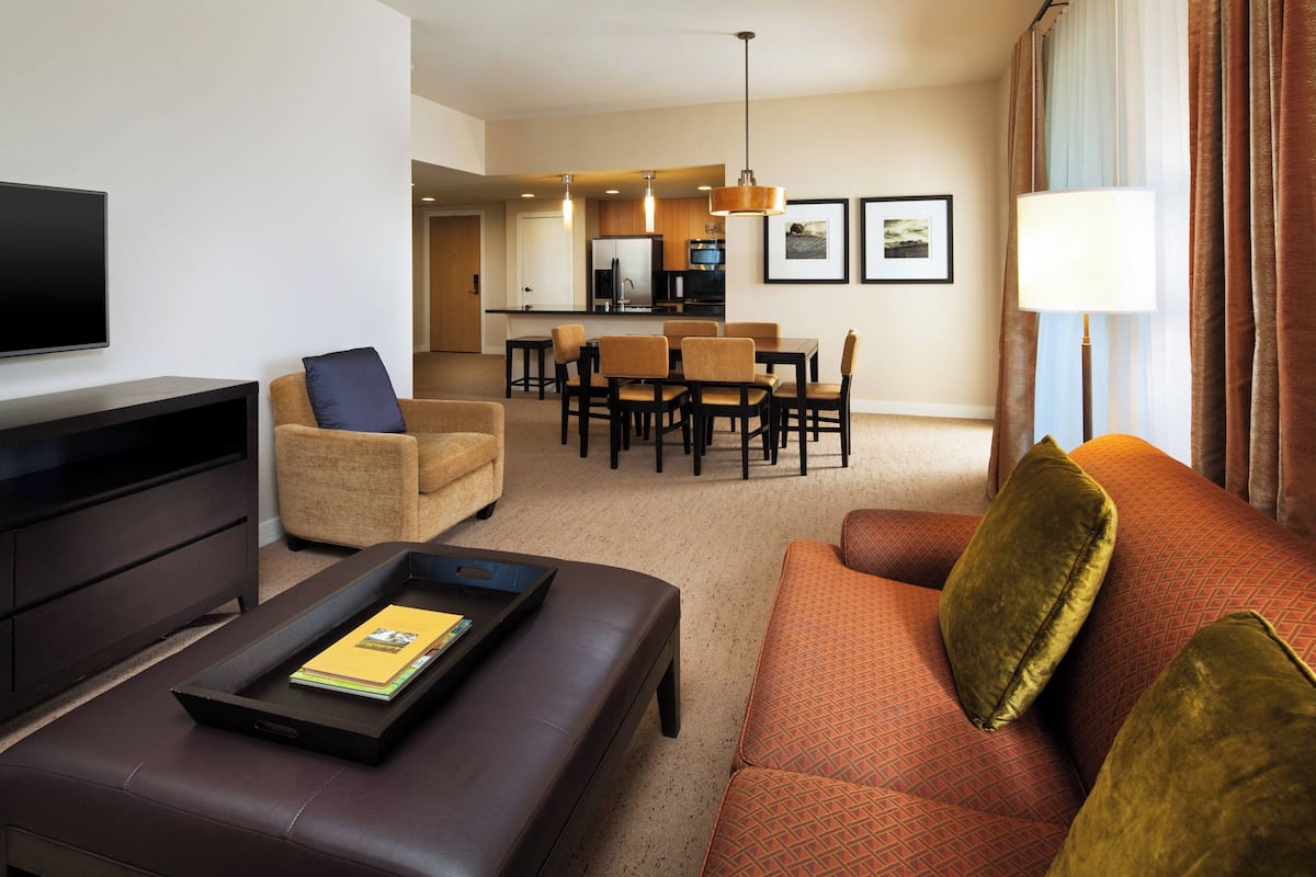 A modern living room with a sofa, a coffee table, and a flat-screen TV. There is a dining area with chairs and a table in the background.