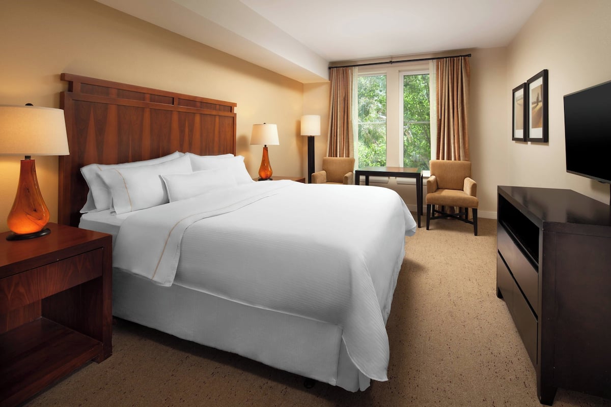 A hotel room with a large bed, two bedside tables with lamps, a small table with a chair, a TV, and a window with curtains.