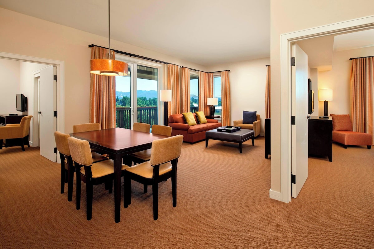 A hotel room with a dining area, a sofa, a TV, and a balcony with curtains.