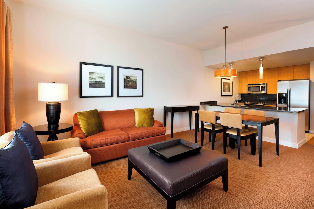 A hotel room with a living area, a dining table, a sofa, and a kitchen area with a refrigerator and microwave.