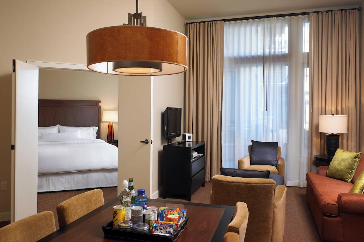 A hotel room with a bed, a TV, a desk with a lamp, and a seating area with a sofa and chairs.