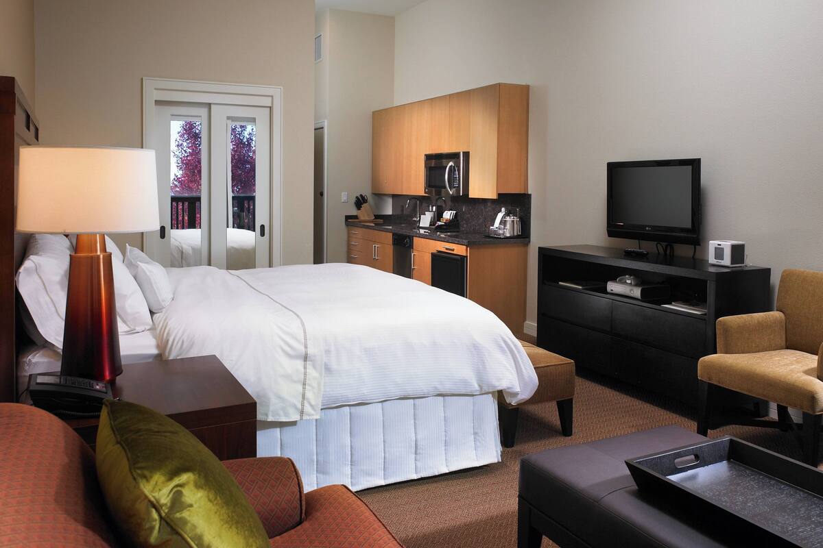 A hotel room with a bed, a desk, a television, a small kitchenette, and a seating area.