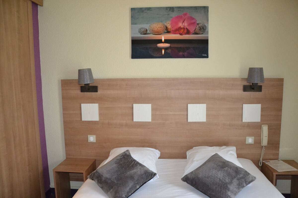 A hotel room with a wooden headboard, two bedside tables with lamps, a bed with white linens and gray pillows, and a painting above the headboard.