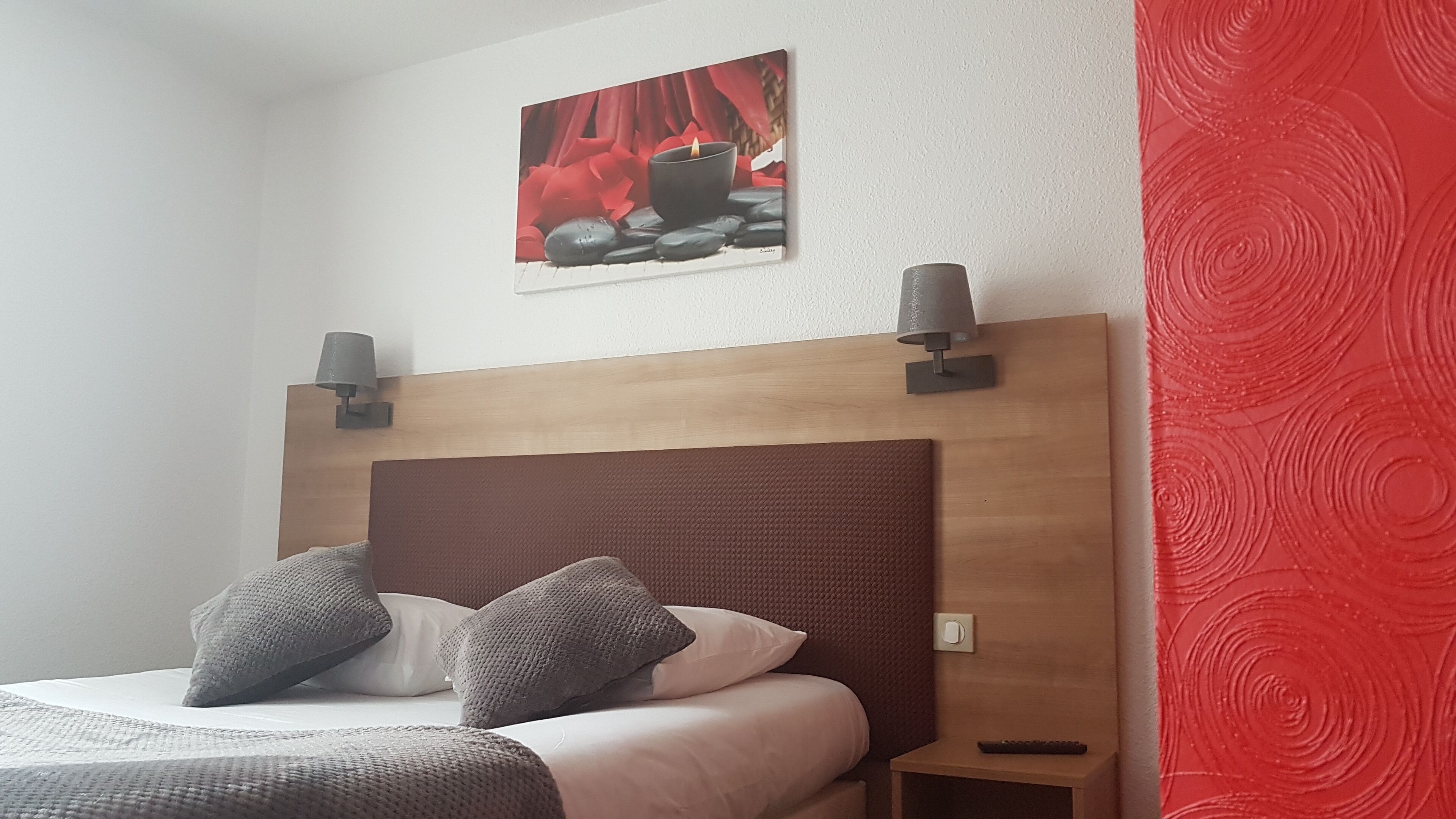 A hotel room with a wooden headboard, two bedside lamps, a painting of red flowers, and a red textured wall.