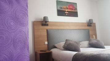 Camera d'albergo con una testiera in legno, due abat-jour, un comodino, un letto con due cuscini e un quadro appeso al muro raffigurante frutta e fiori.