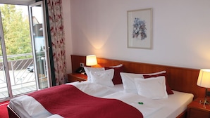 Una habitación de hotel con una cama grande, una mesita de noche con lámpara, un cuadro enmarcado en la pared y un ventanal corredero al exterior.