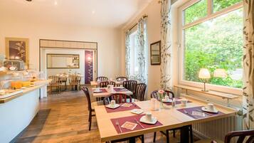 A dining area with tables set for breakfast, a kitchen in the background, and large windows with curtains.