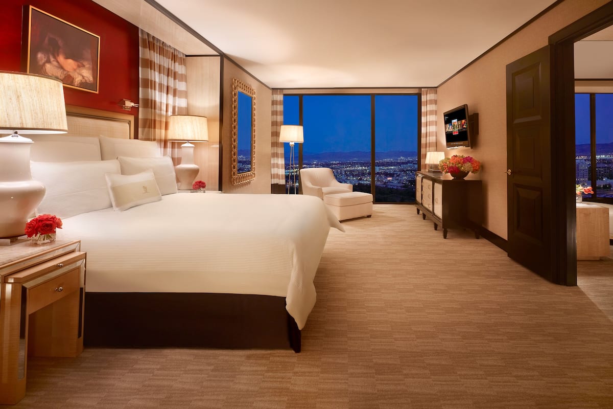 A spacious hotel room with a large bed, a sitting area, a desk, and a view of the city at night.