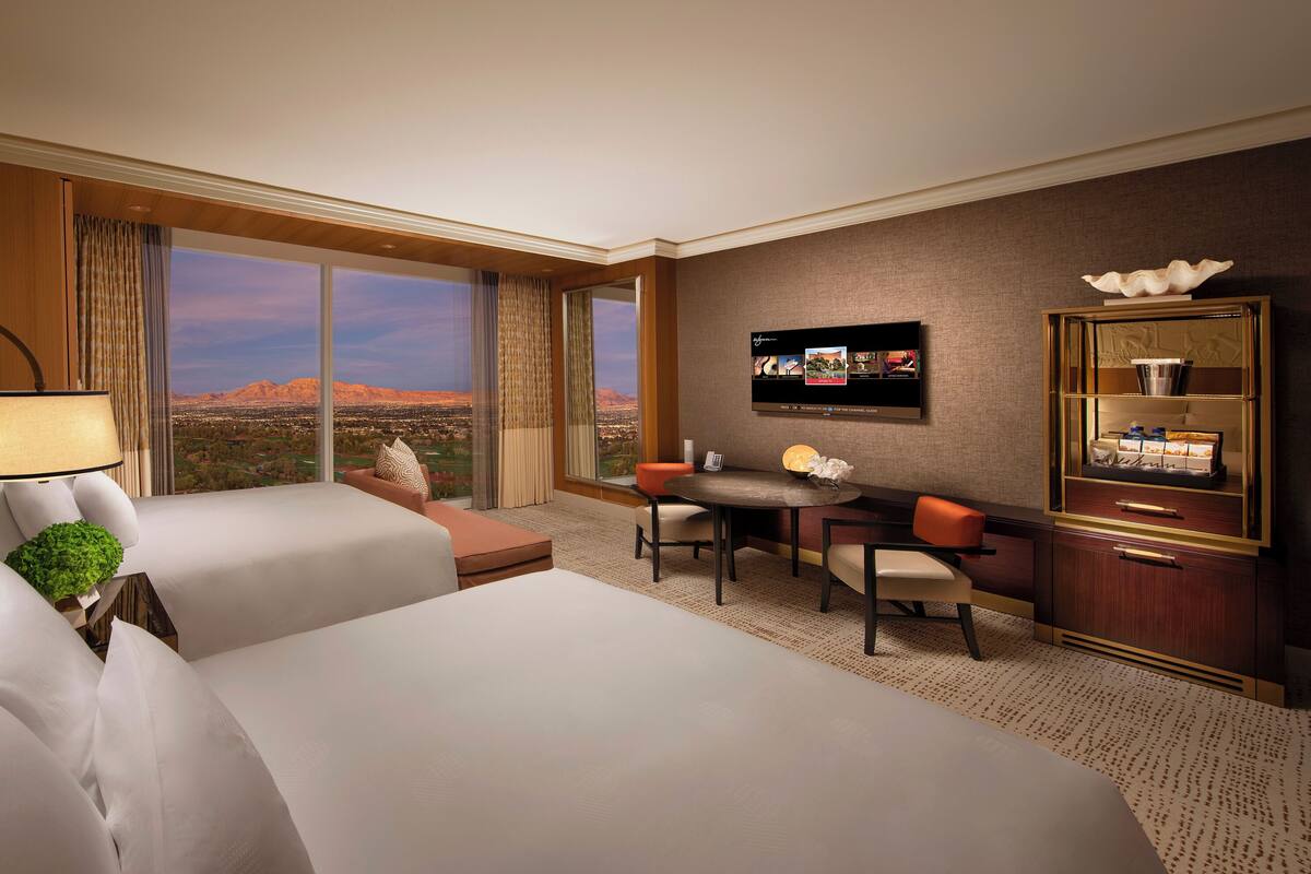 A hotel room with two beds, a desk with a chair, a television, and a view of the desert.