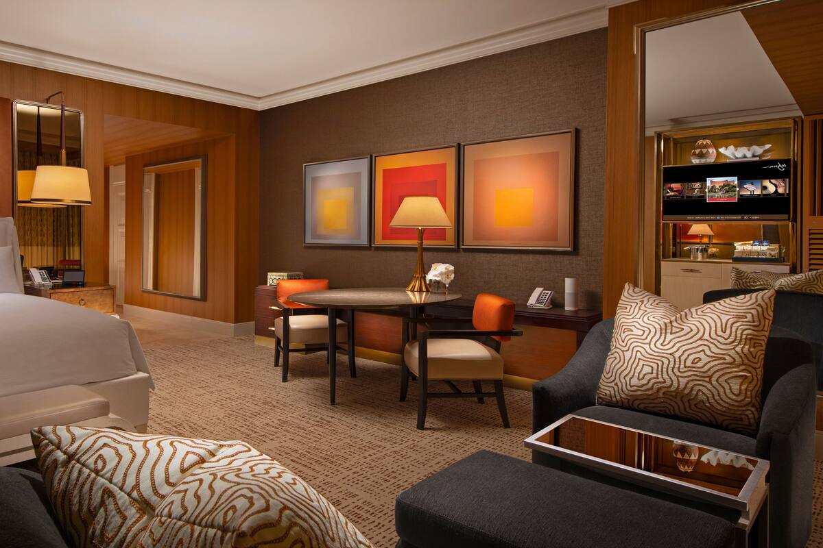 A hotel room with a large bed, a desk with a chair, a sofa, and a small table. There are framed pictures on the wall and a lamp on the desk.