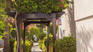 Un sendero en un jardín, con un pórtico de madera, rodeado de abundante follaje y plantas florecientes.