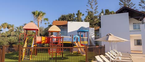 A playground with slides and climbing structures, surrounded by a fence, and a pool area with lounge chairs and umbrellas.