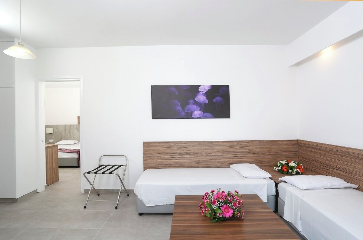 A hotel room with a wooden headboard, two single beds, a small table with a vase of flowers, a wall-mounted picture, and a metal drying rack.