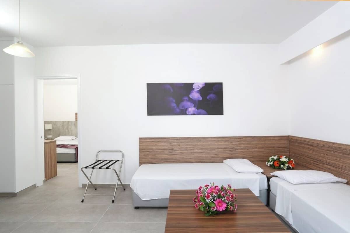 A hotel room with a wooden headboard, two single beds, a small table with a vase of flowers, a wall-mounted picture, and a metal drying rack.