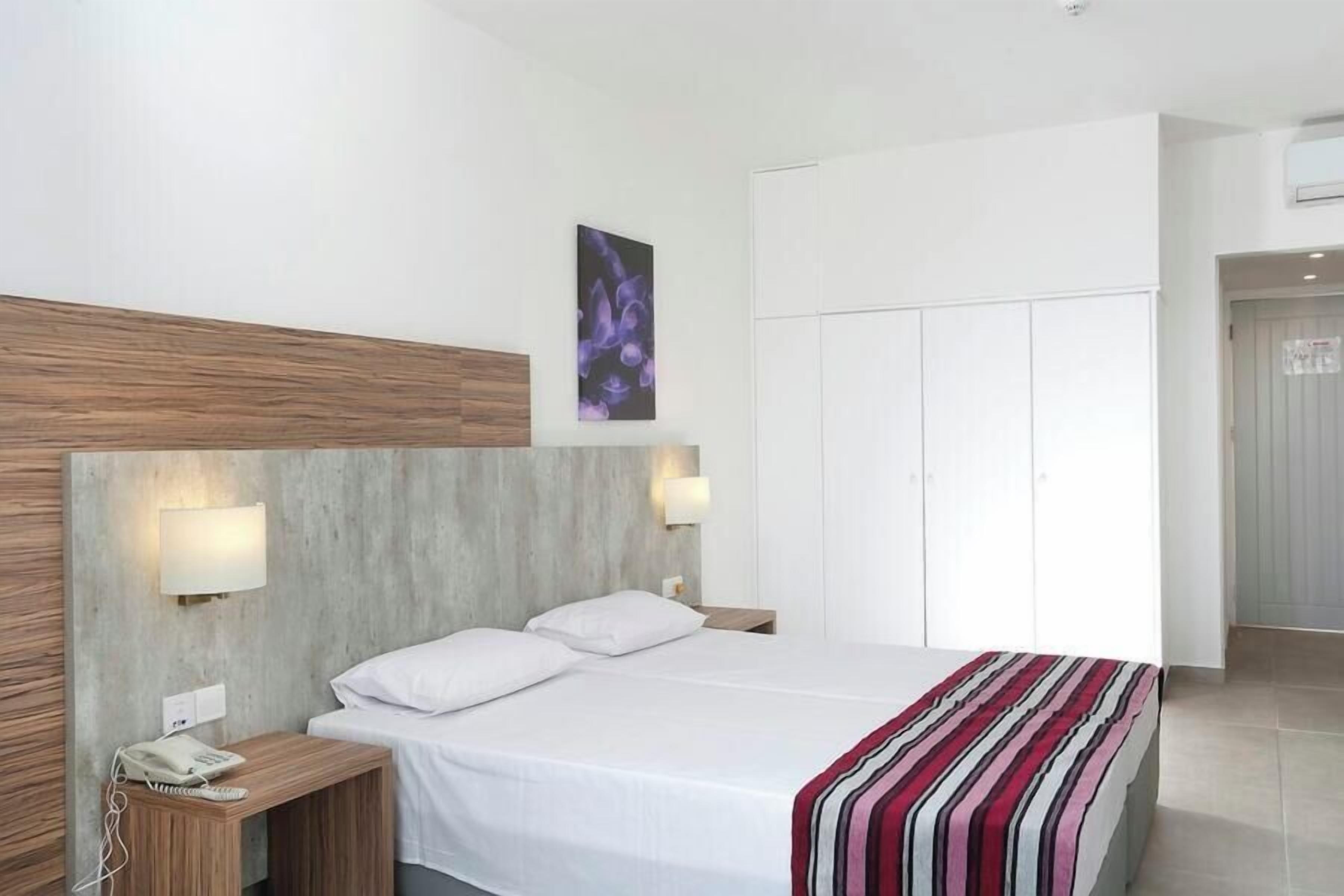 A hotel room with a bed, bedside tables, a phone, a wall-mounted light, a framed picture, and a wardrobe.