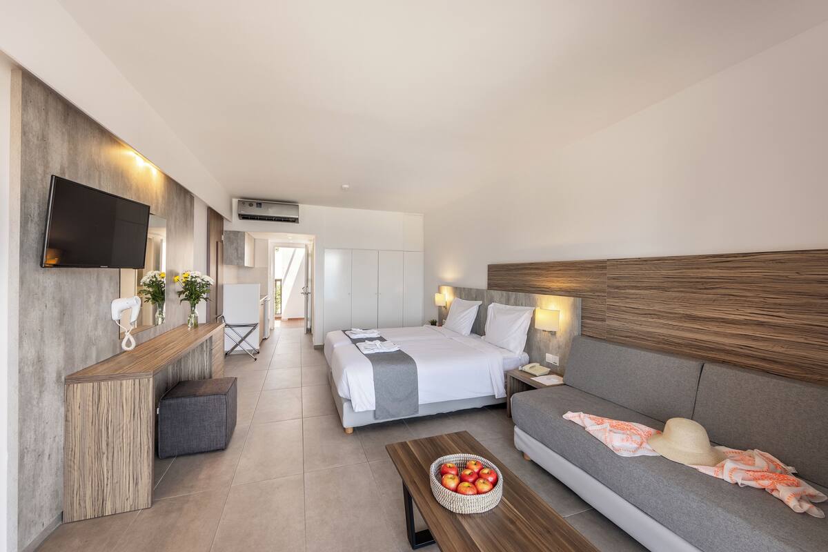 A modern hotel room with a bed, sofa, TV, and a small table with fruit.