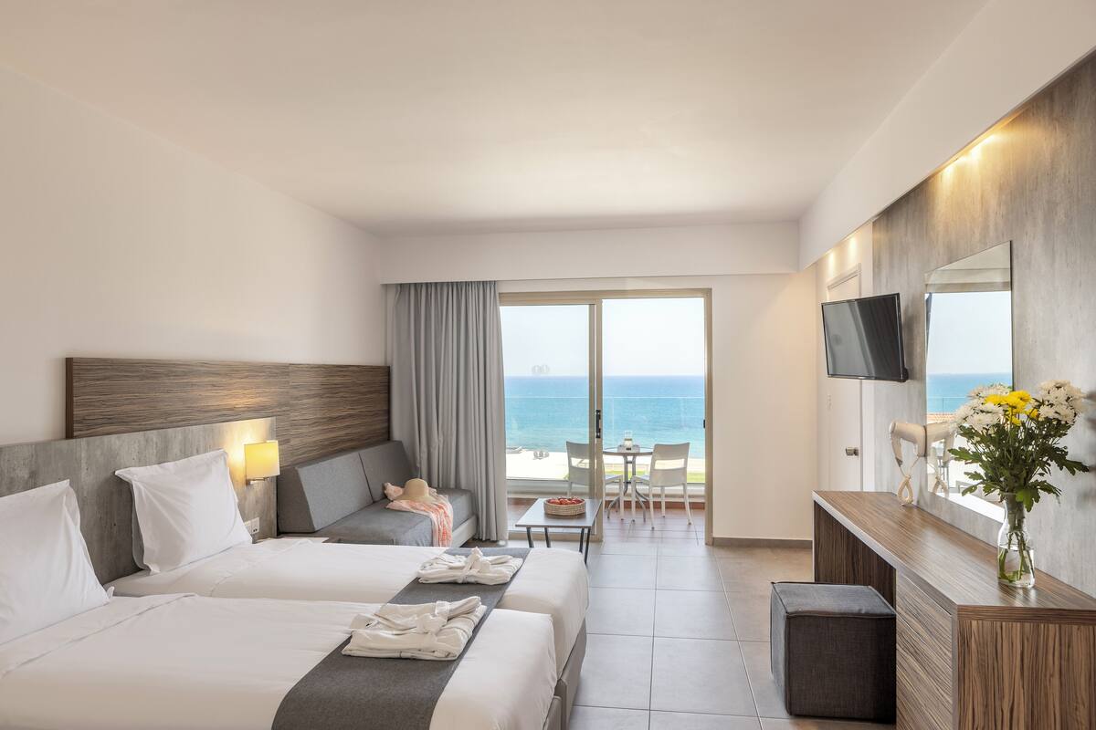 A hotel room with two beds, a sofa, a TV, a dining table with chairs, and a view of the ocean.
