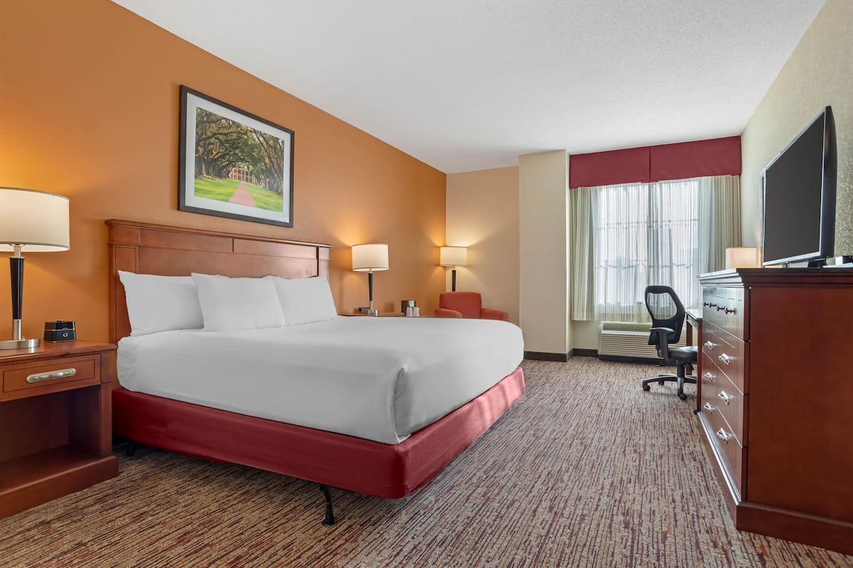A hotel room with a large bed, two bedside tables with lamps, a desk with a chair, a television, and a window with curtains.