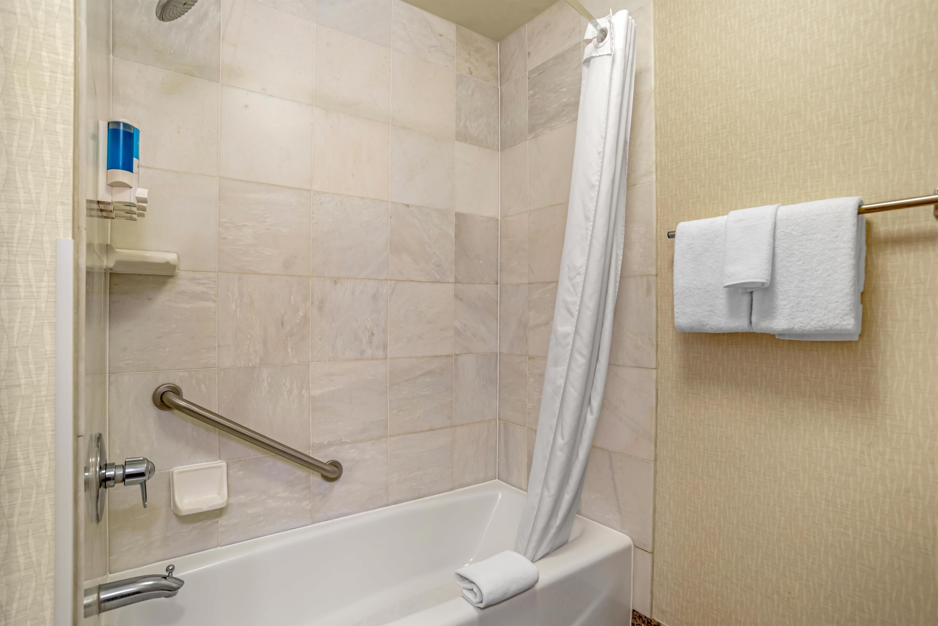 A bathroom with a white bathtub, a shower curtain, a towel rack with two towels, a soap dispenser, and a handrail.
