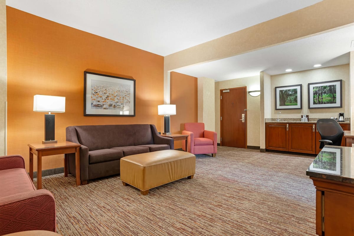 A hotel lobby with a brown sofa, a pink chair, a wooden table, and a kitchenette area.