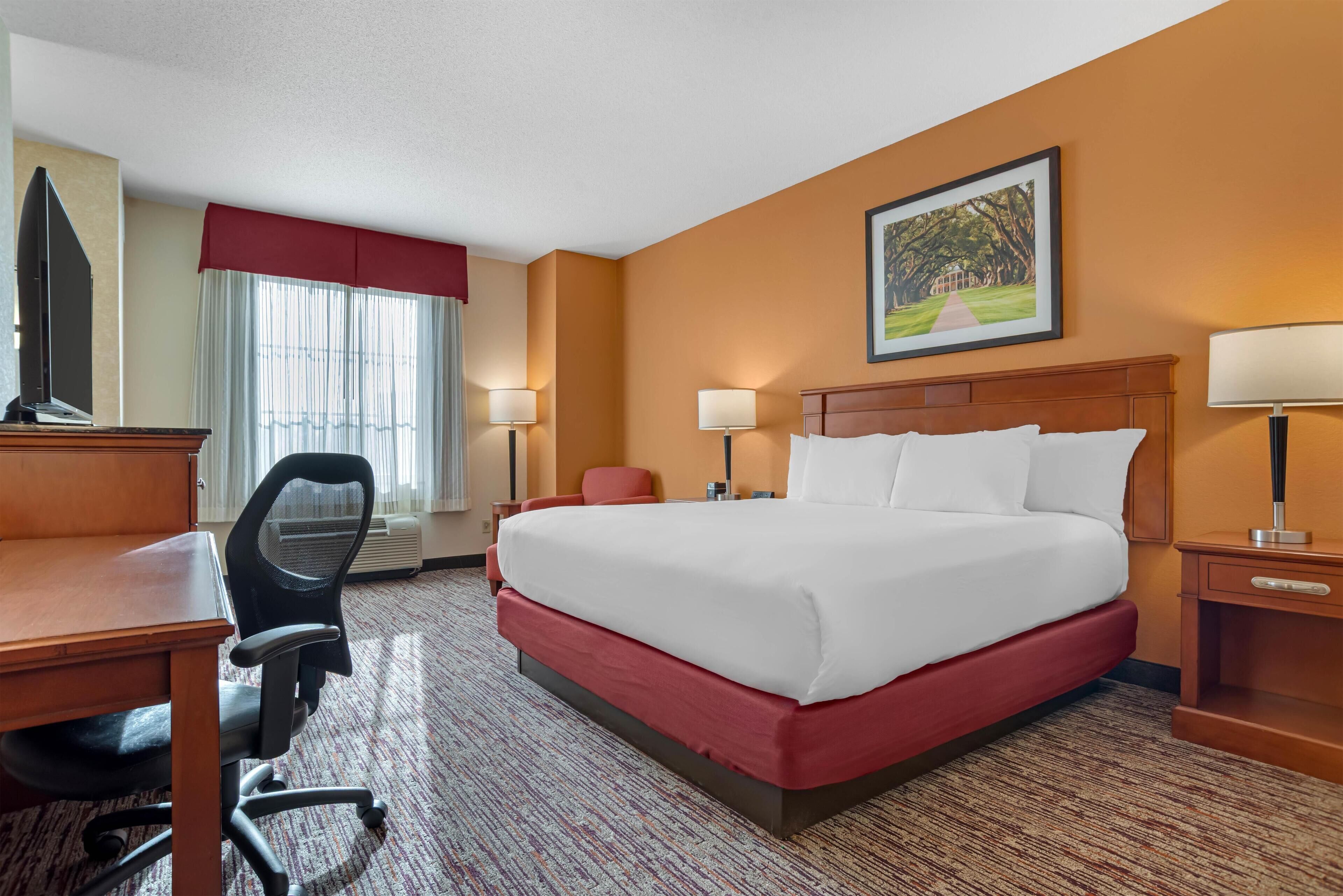 A hotel room with a large bed, a desk with a chair, two bedside lamps, a TV, and a window with curtains.