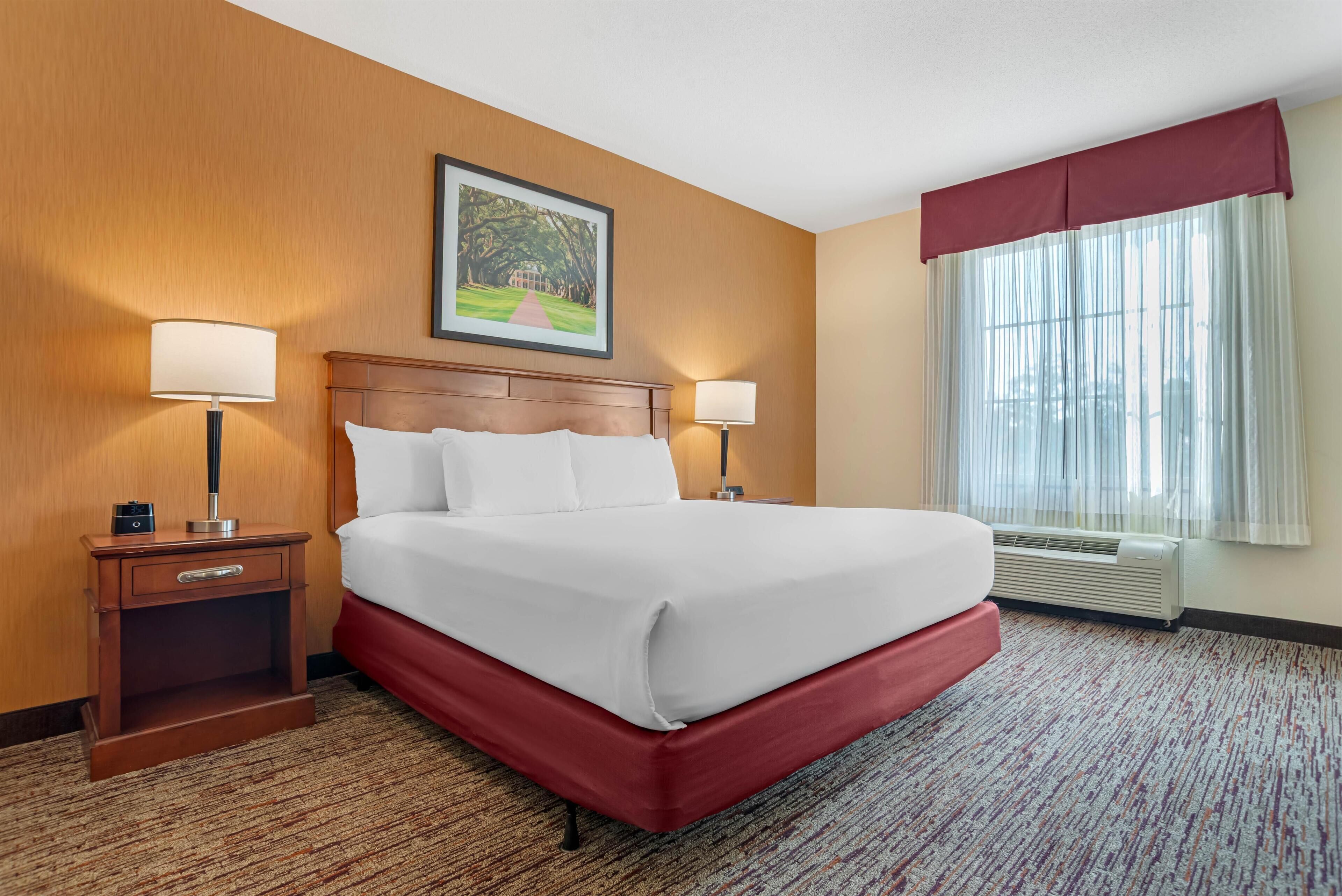 Una habitación de hotel con una cama grande, dos mesitas de noche, una lámpara de noche, un cuadro enmarcado en la pared y una ventana con cortinas.