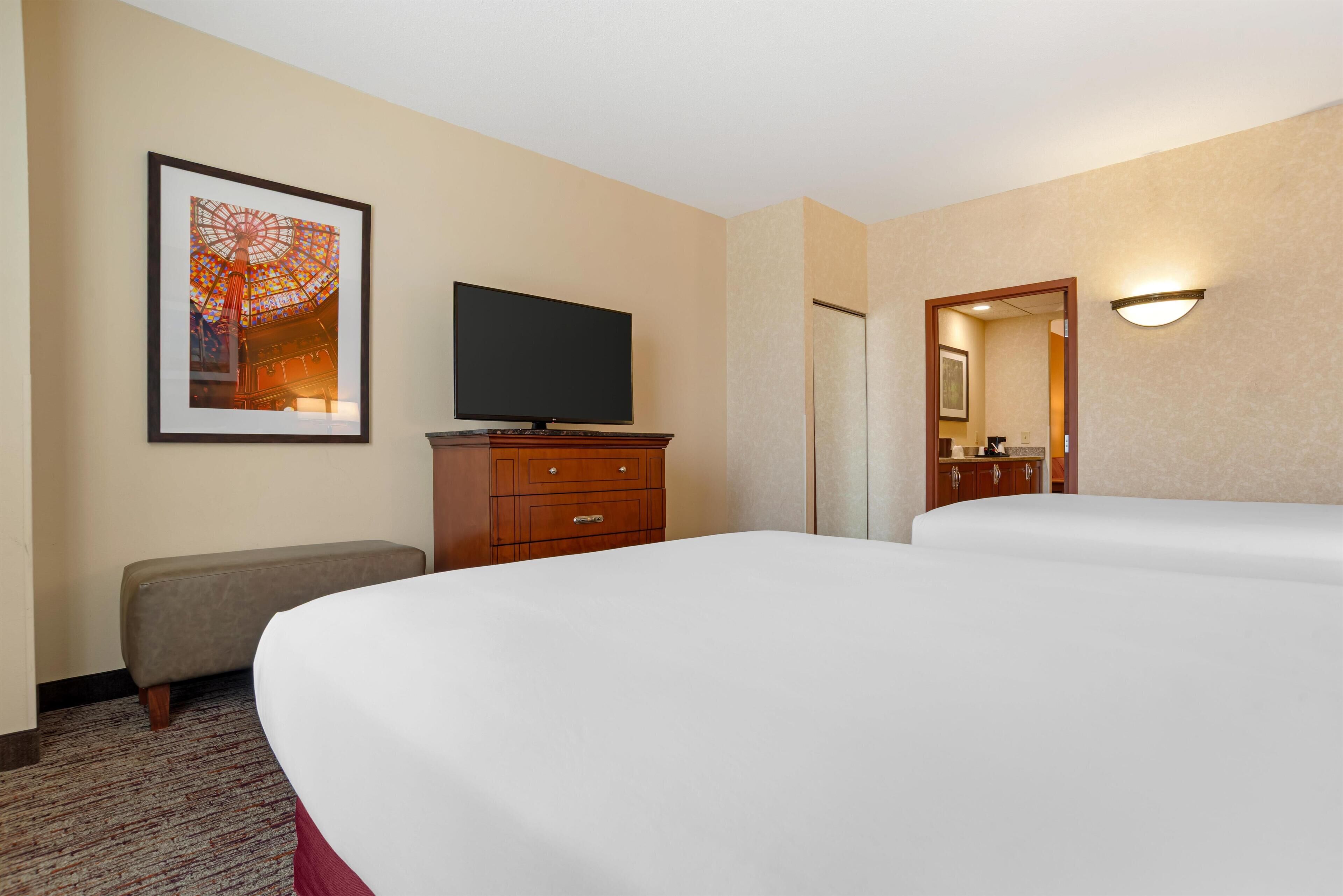 Hotelkamer met twee bedden, een flatscreen televisie, een houten dressoir, een bankje, een ingelijste foto aan de muur en een spiegel.