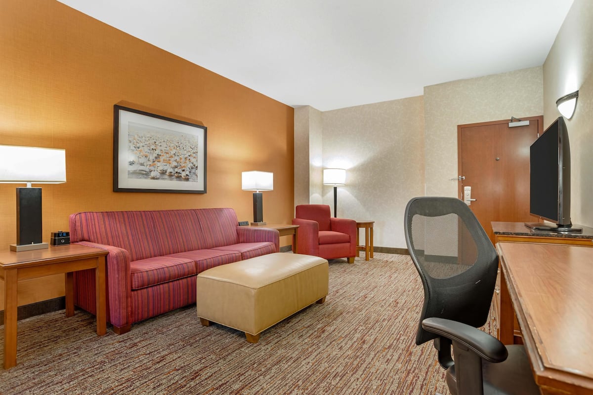 A hotel room with a red-striped sofa, a beige ottoman, a red armchair, a black office chair, a wooden desk, and a television.