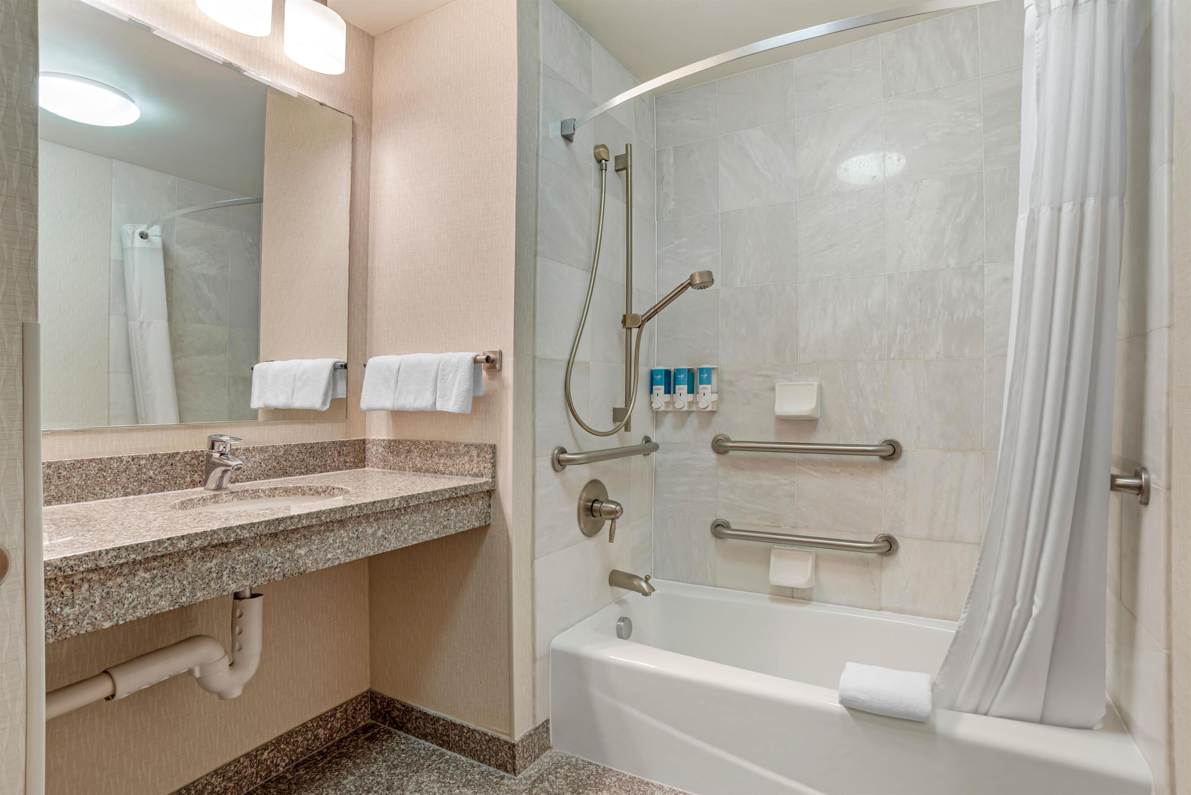 A modern bathroom with a granite countertop sink, a bathtub, and a shower area with grab bars.