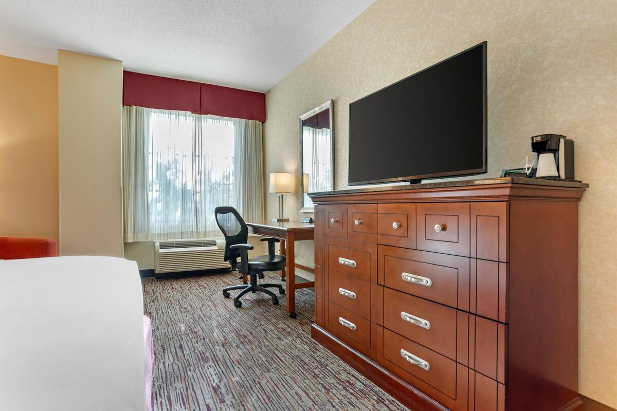 A hotel room with a bed, a desk with a chair, a television, a dresser, and a window with curtains.