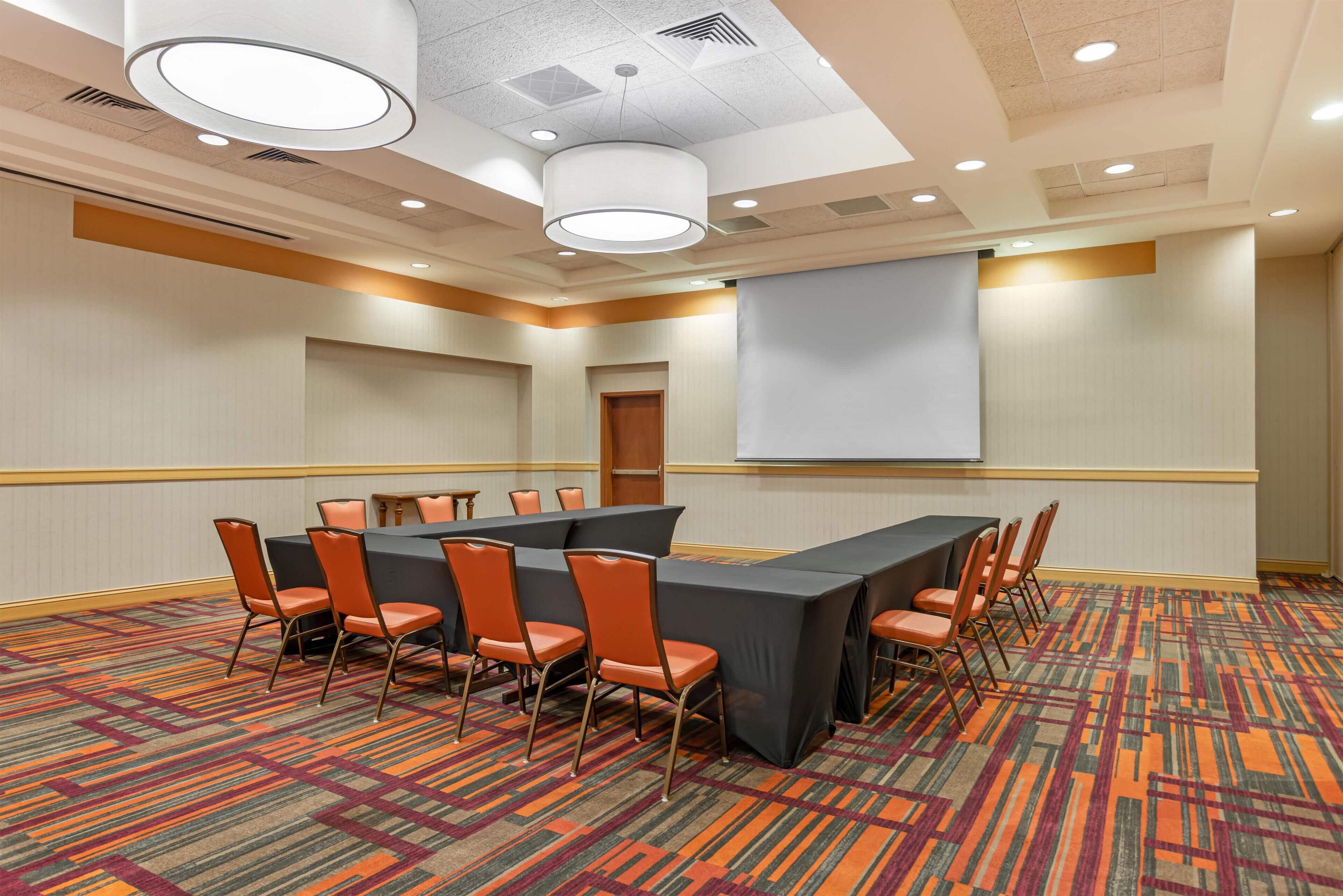A conference room with a long table, chairs, a projector screen, and a carpeted floor.