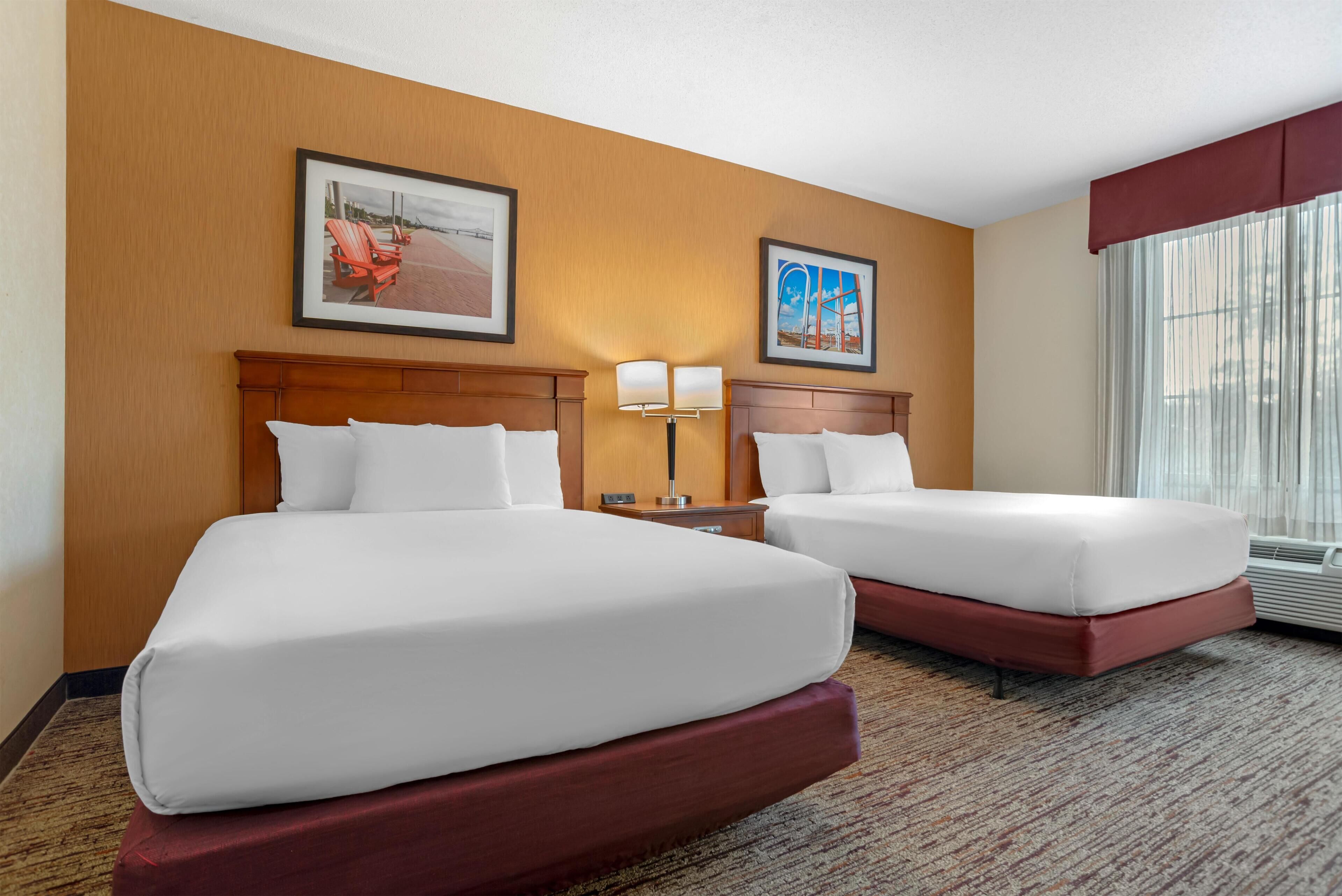 Quarto de hotel com duas camas, cabeceira de madeira, mesa de cabeceira com abajur e duas quadros na parede.