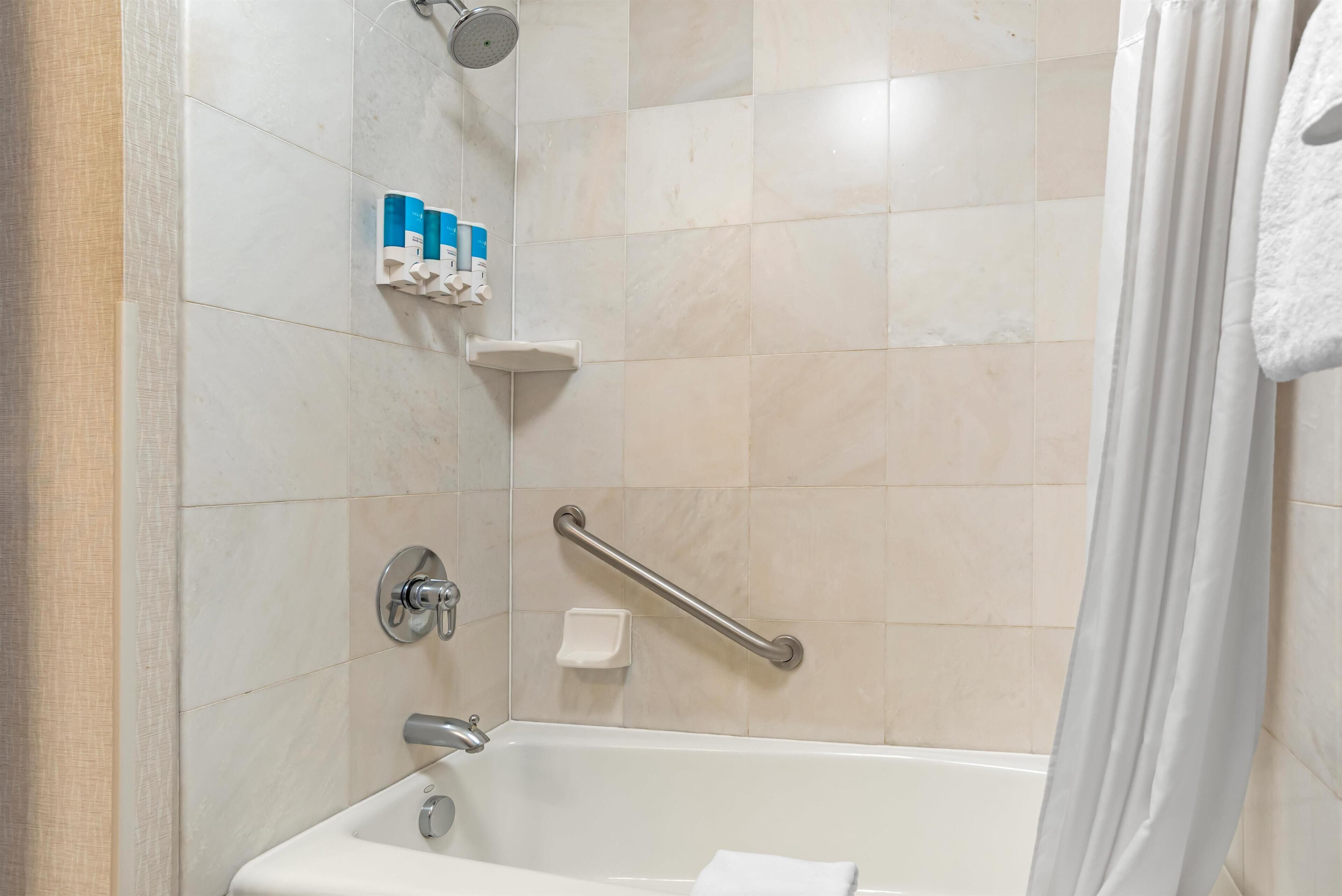 A bathroom with a bathtub, a showerhead, a handrail, and toiletries on a shelf.
