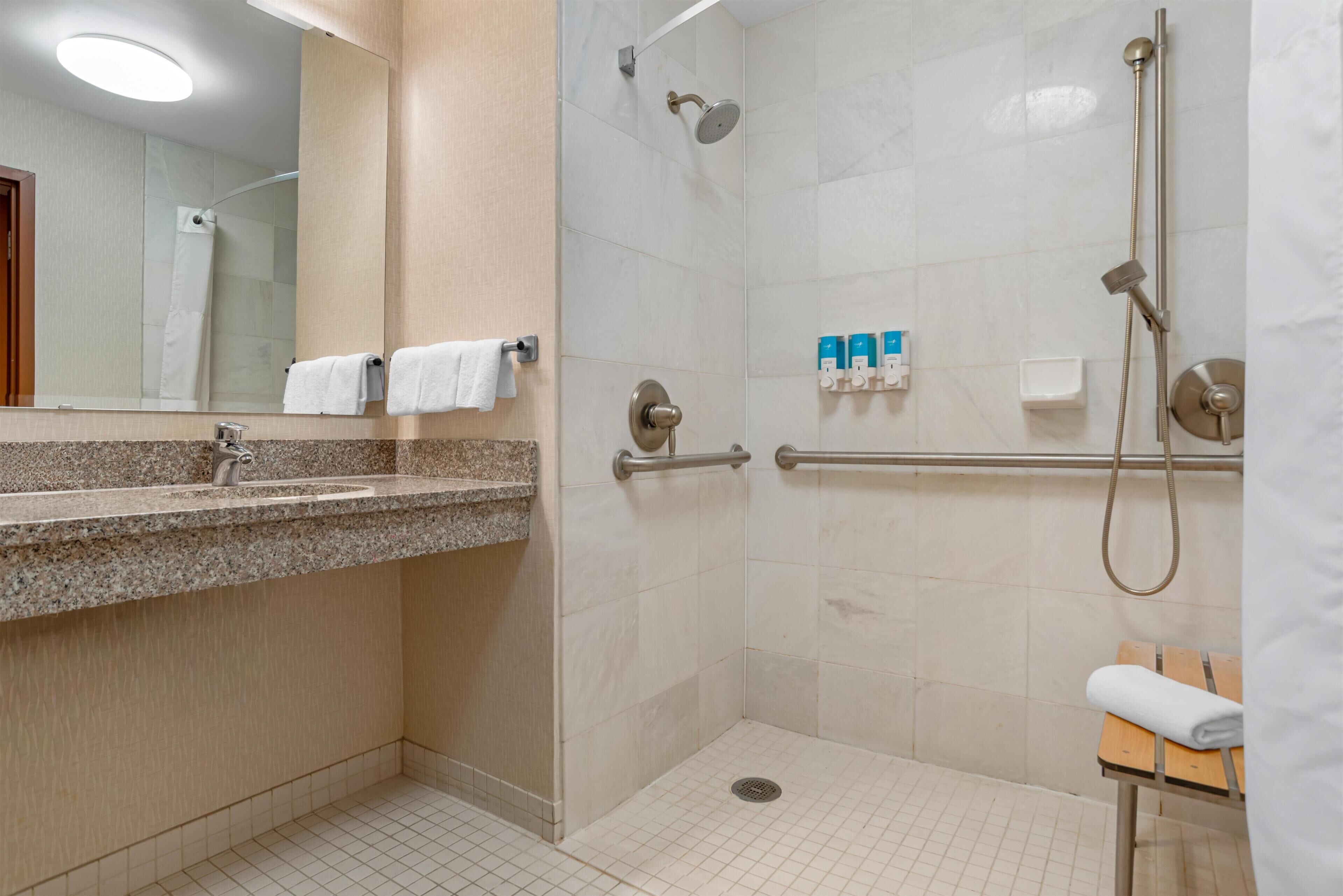 A modern bathroom with a granite countertop sink, a glass shower enclosure, and a small wooden bench.