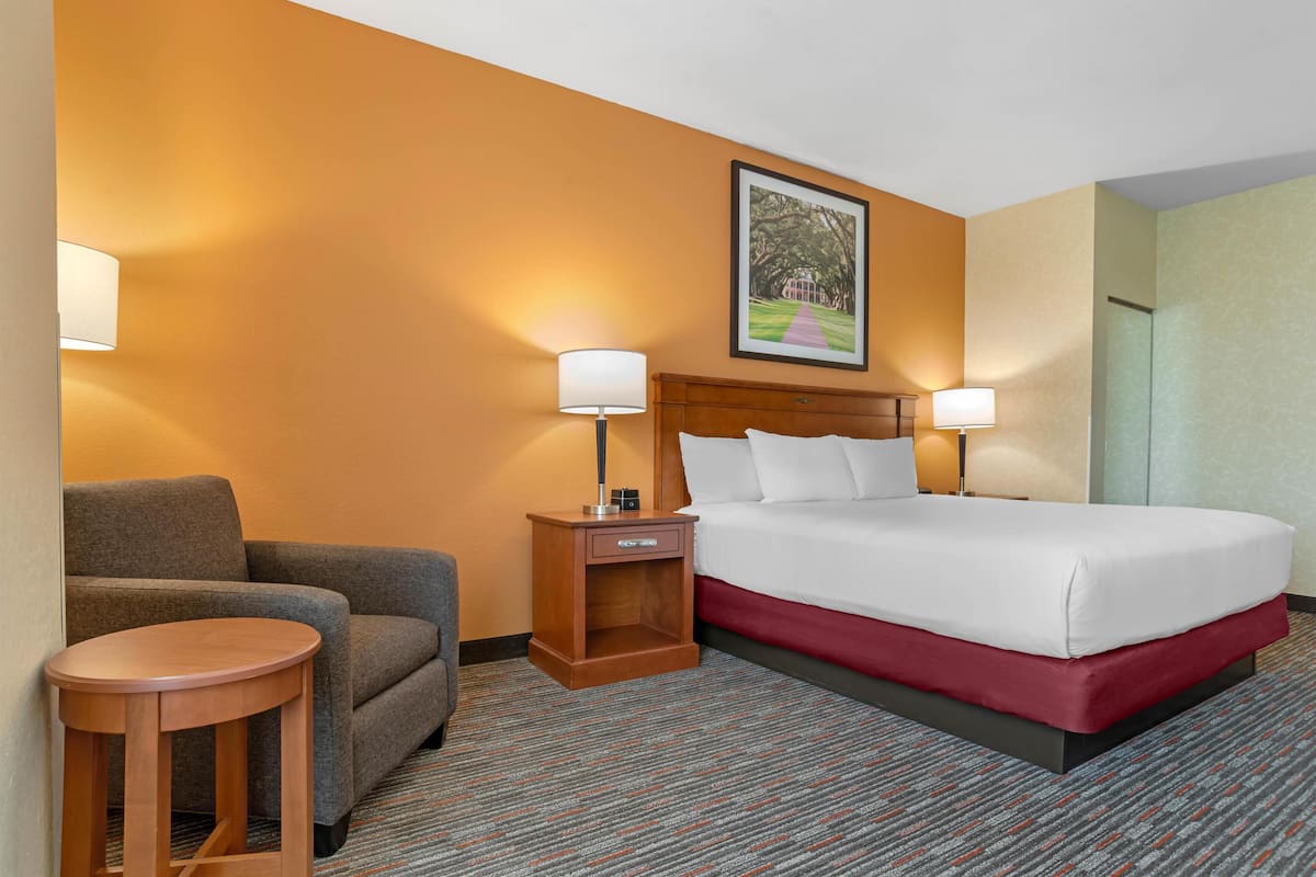 A hotel room with a bed, a chair, a small table, and a nightstand with lamps.