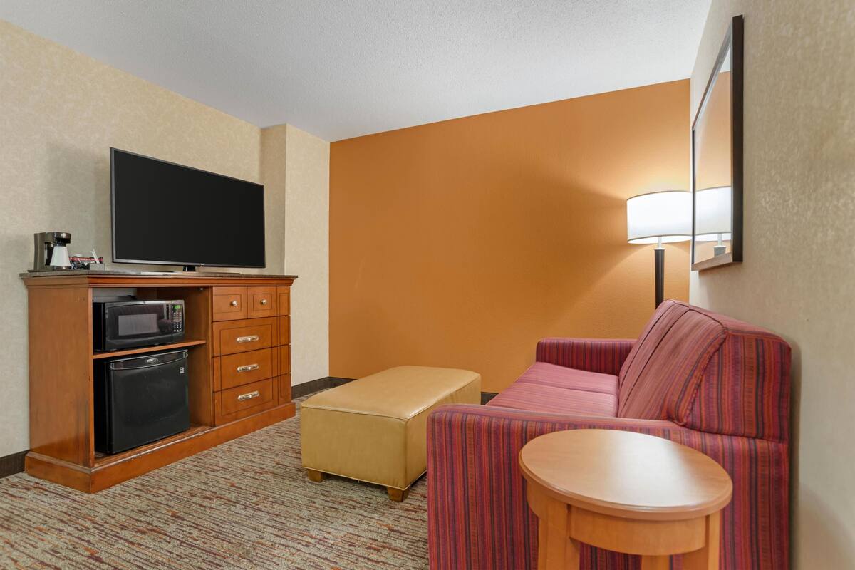 A hotel room with a flat-screen TV, a microwave, a refrigerator, a sofa, an ottoman, and a small wooden table.