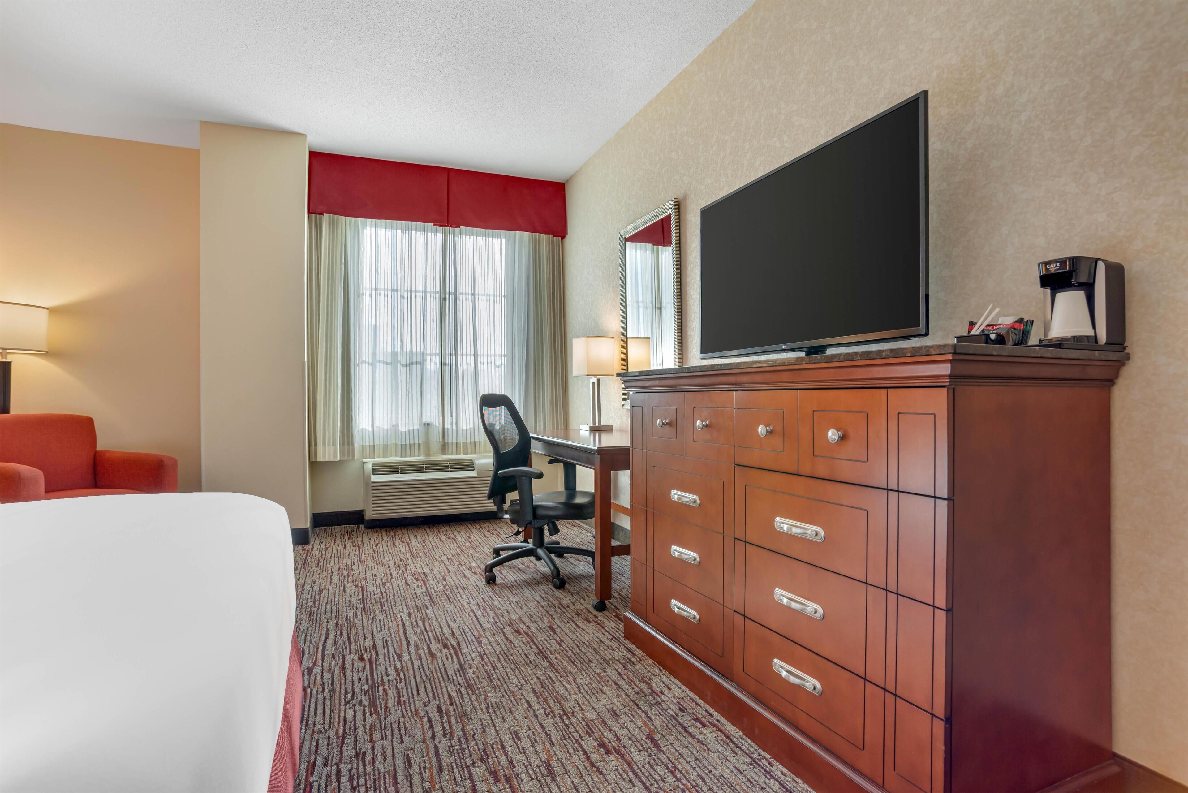 A hotel room with a bed, a desk with a chair, a television, a dresser, and a window with curtains.