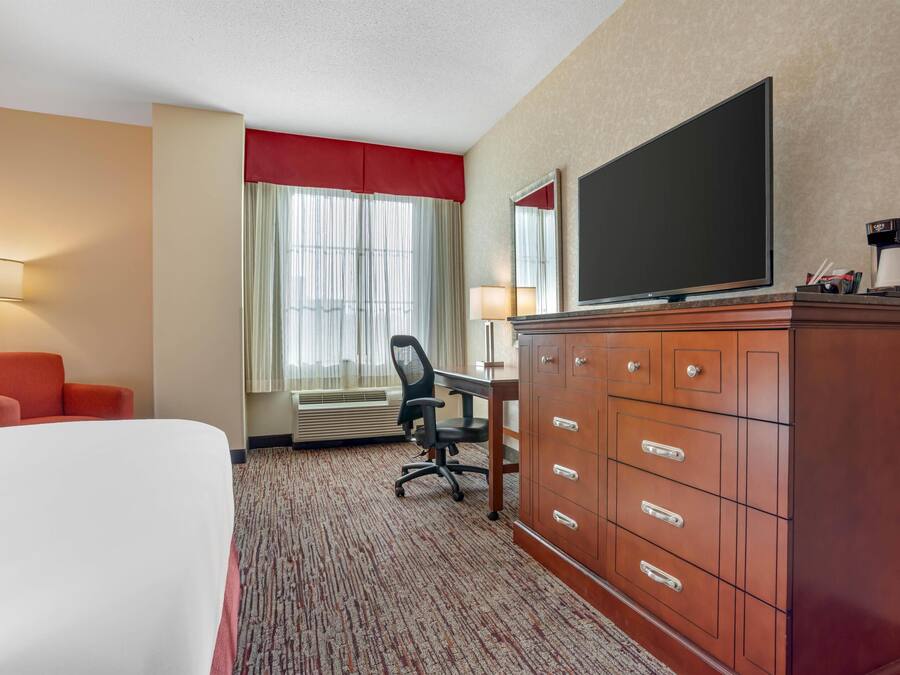 A hotel room with a bed, a desk with a chair, a television, a dresser, and a window with curtains.