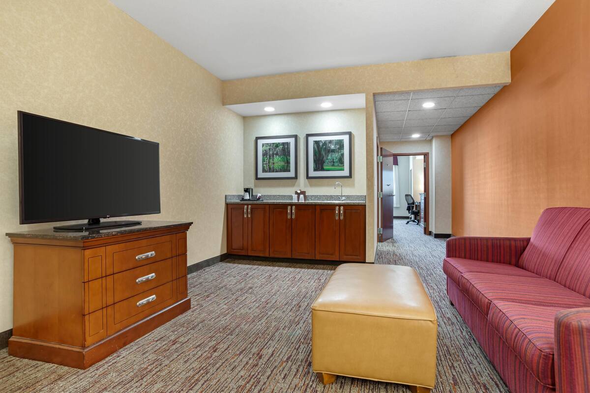 A hotel room with a flat-screen TV, wooden cabinet, kitchenette, and a red-striped sofa.