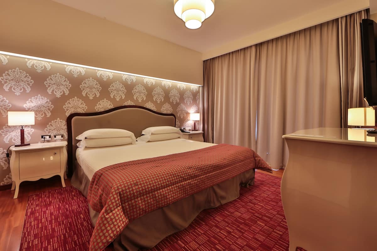 Une chambre d’hôtel avec un grand lit, deux tables de chevet avec des lampes, un bureau avec une chaise, un grand miroir et une télévision.