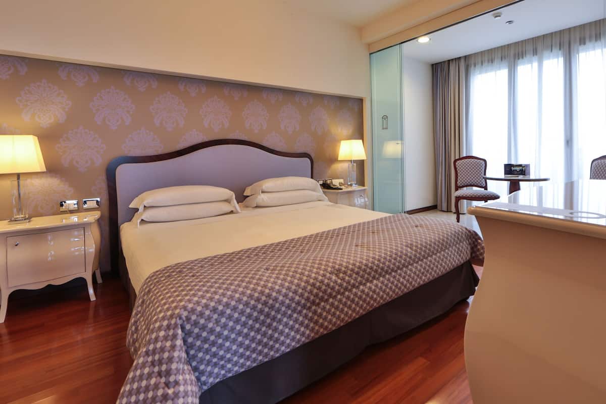 Une chambre d’hôtel avec un grand lit, deux tables de chevet avec des lampes, une petite table avec une chaise et une grande fenêtre avec des rideaux.