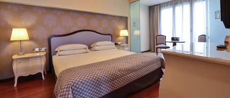A hotel room with a large bed, two bedside tables with lamps, a small table with a chair, and a large window with curtains.