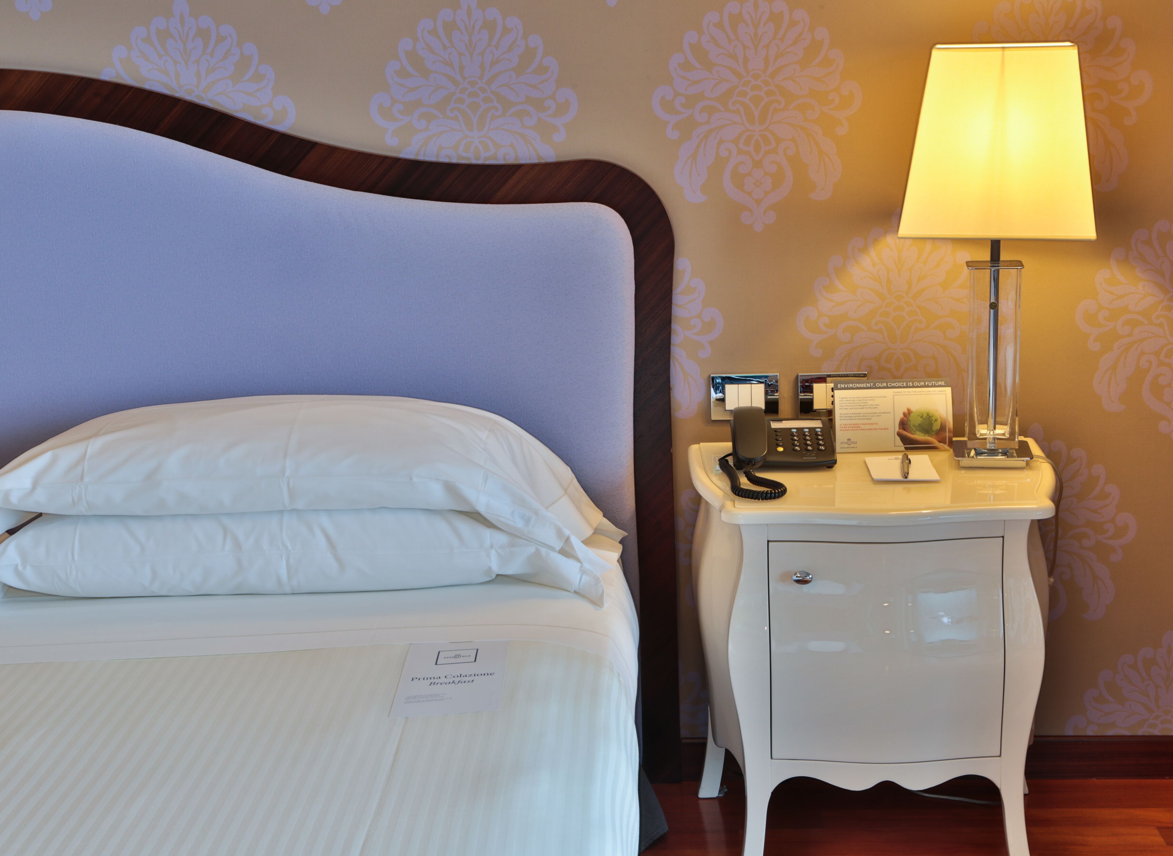 A neatly made bed with pillows, a bedside table with a lamp, a phone, and a sign indicating breakfast availability.