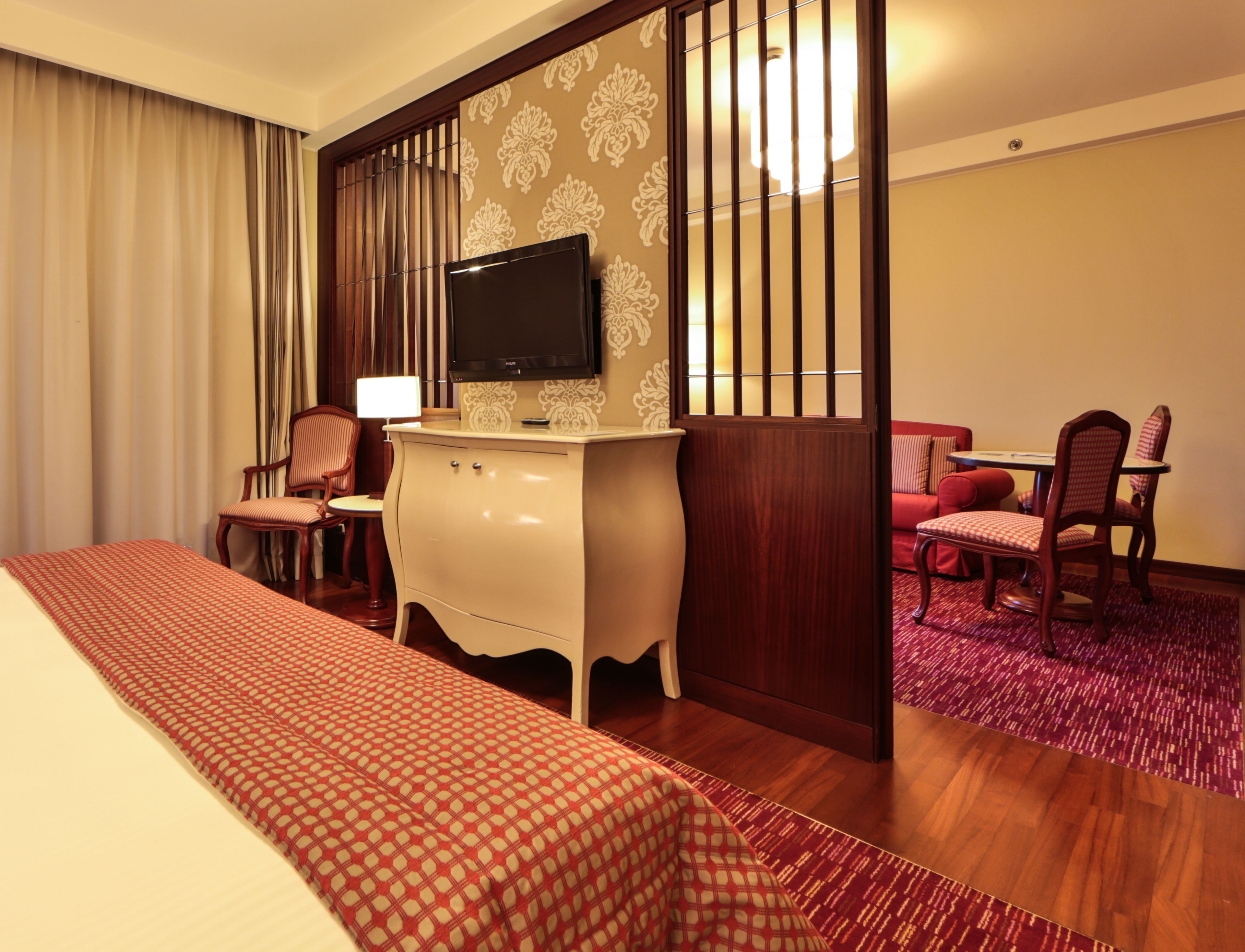 Una habitación de hotel con una cama, un escritorio con lámpara, un televisor, un sofá rojo y una mesa de comedor con sillas.