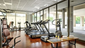 A modern gym with various exercise equipment, large windows, and a wooden table with towels and water bottles. - UNA Hotels Expo Fiera Milano (Pero)