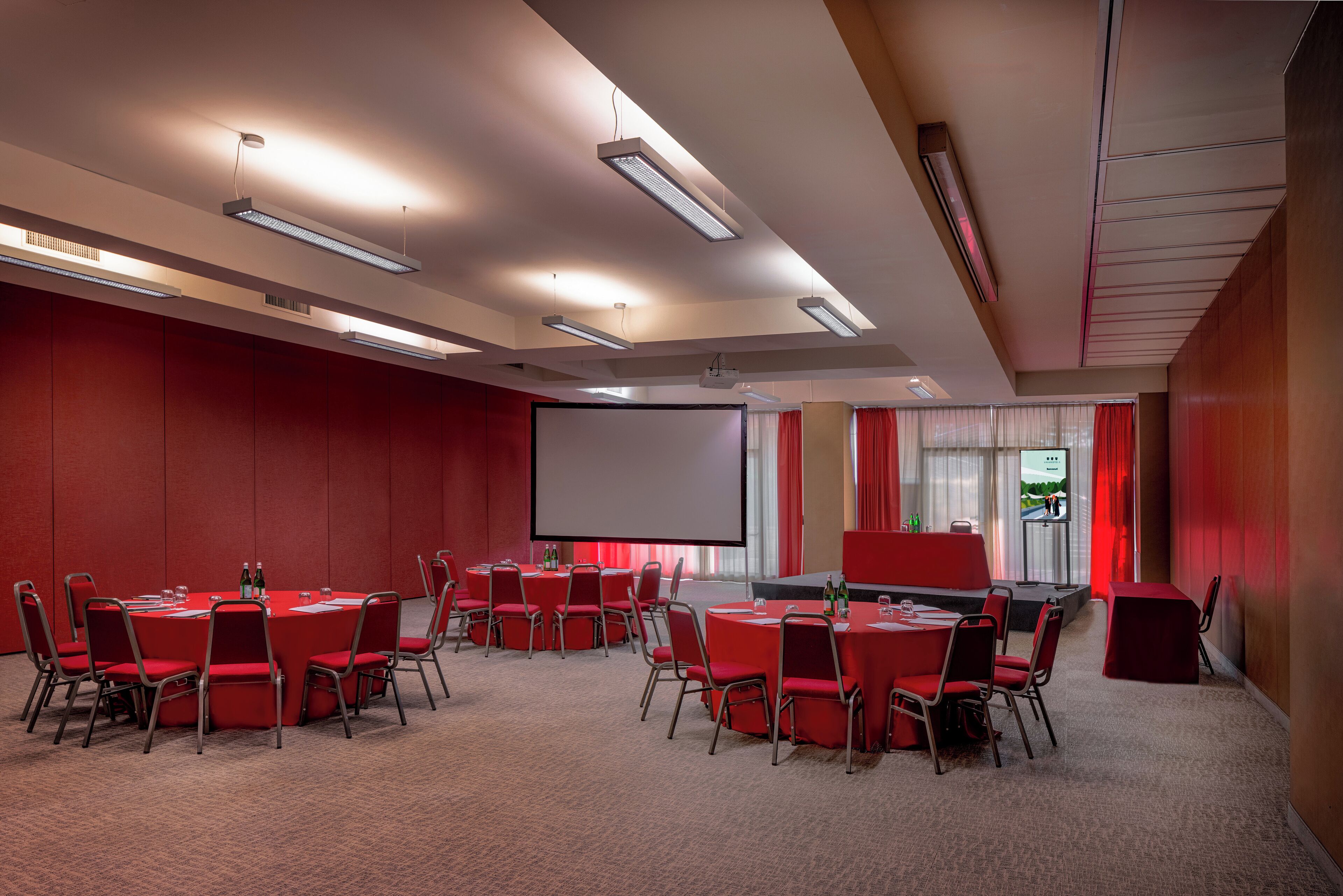 A conference room with multiple tables and chairs, a large screen, and ceiling lights.