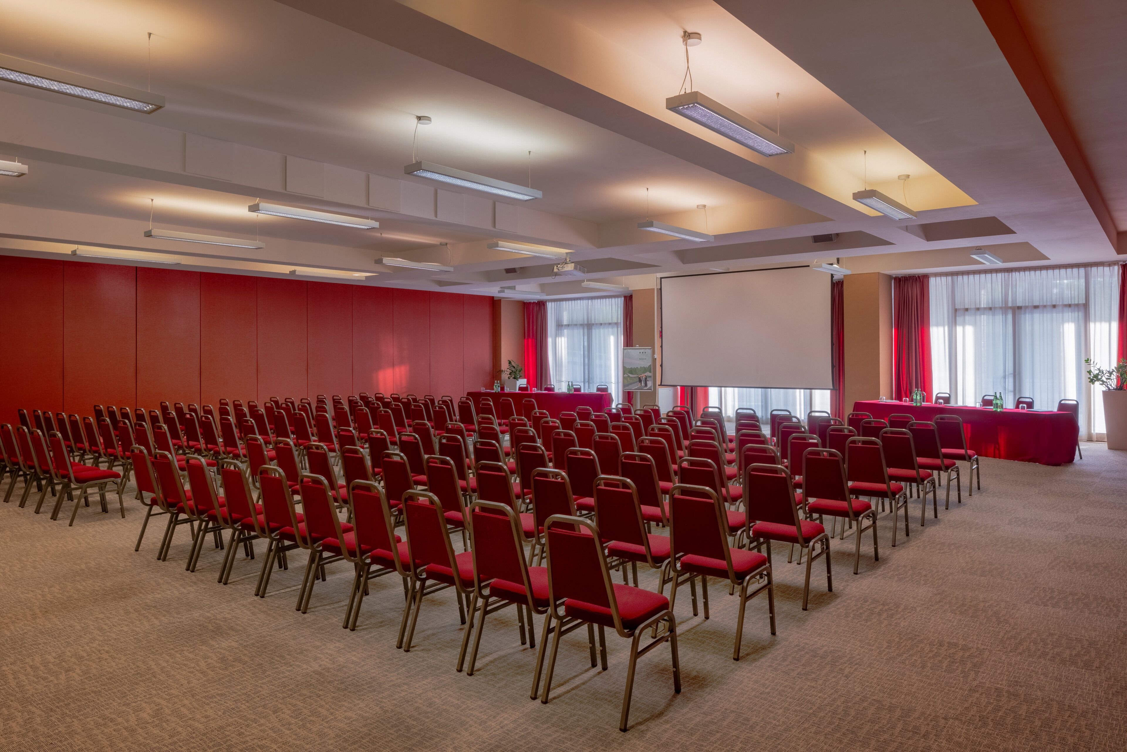 Ein Konferenzraum mit Reihen roter Stühle, einer Projektionsleinwand und einem Tisch mit roter Tischdecke.