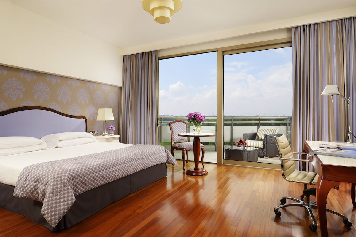 Une chambre d’hôtel avec un grand lit, un bureau, une chaise, une petite table avec des fleurs et un balcon avec vue.