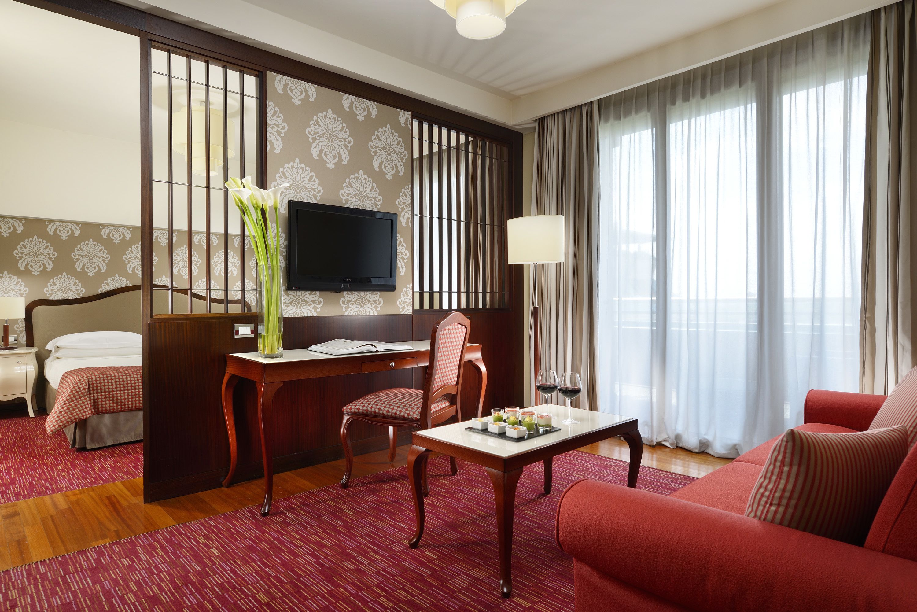 Une chambre d’hôtel avec un lit, un bureau avec une télévision, un canapé rouge et une table à manger avec un tapis rouge.