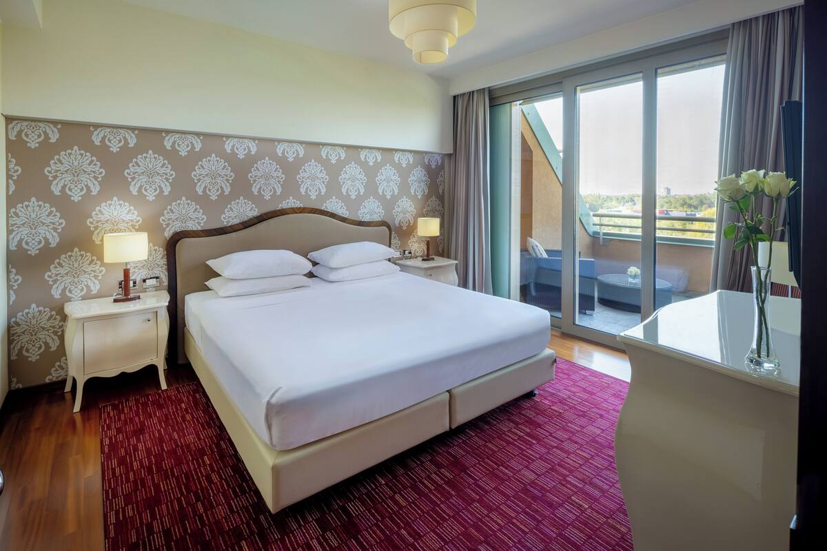Une chambre d’hôtel avec un grand lit, deux tables de chevet avec des lampes, une moquette rouge, une grande fenêtre donnant sur l’extérieur et un vase rempli de fleurs posé sur une table.